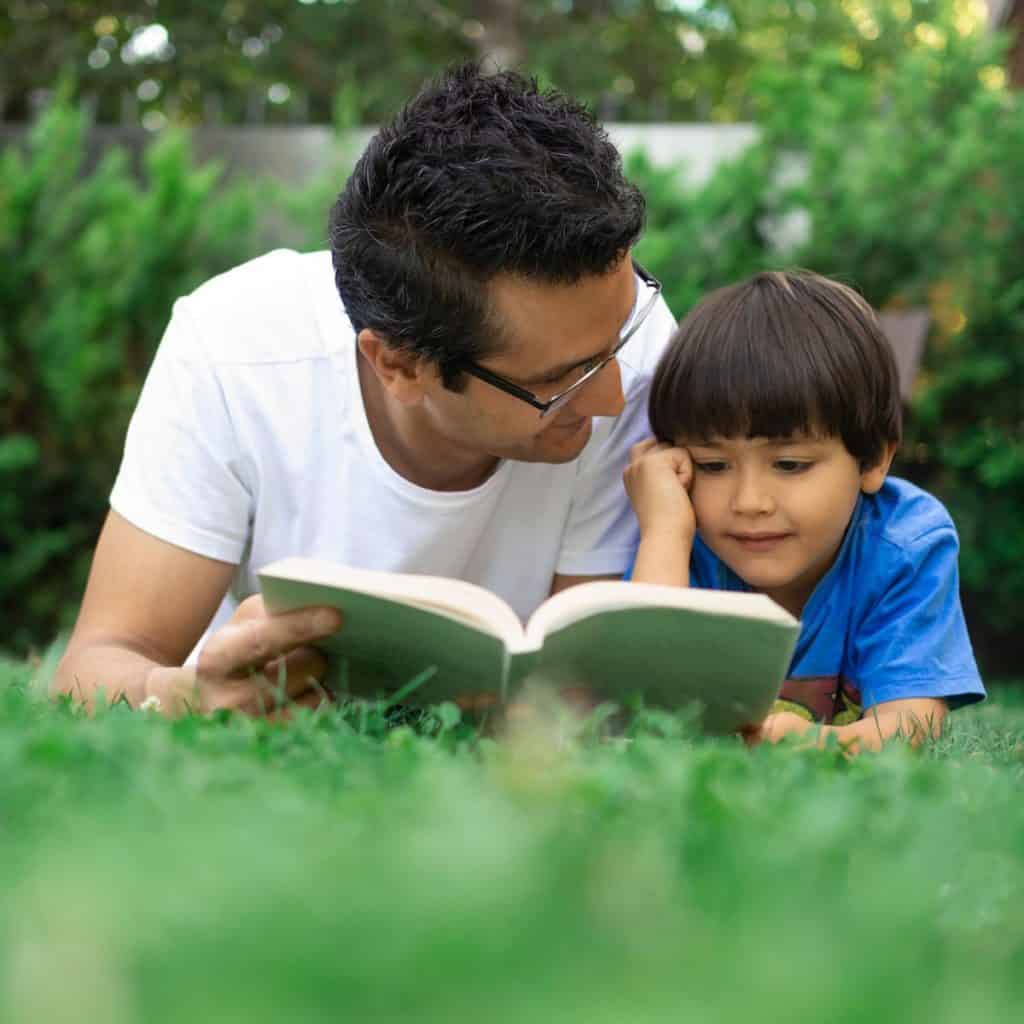 Photo of an adult male in a white shirt and a young boy in a blue shirt laying down in the grass on their stomach reading a book.